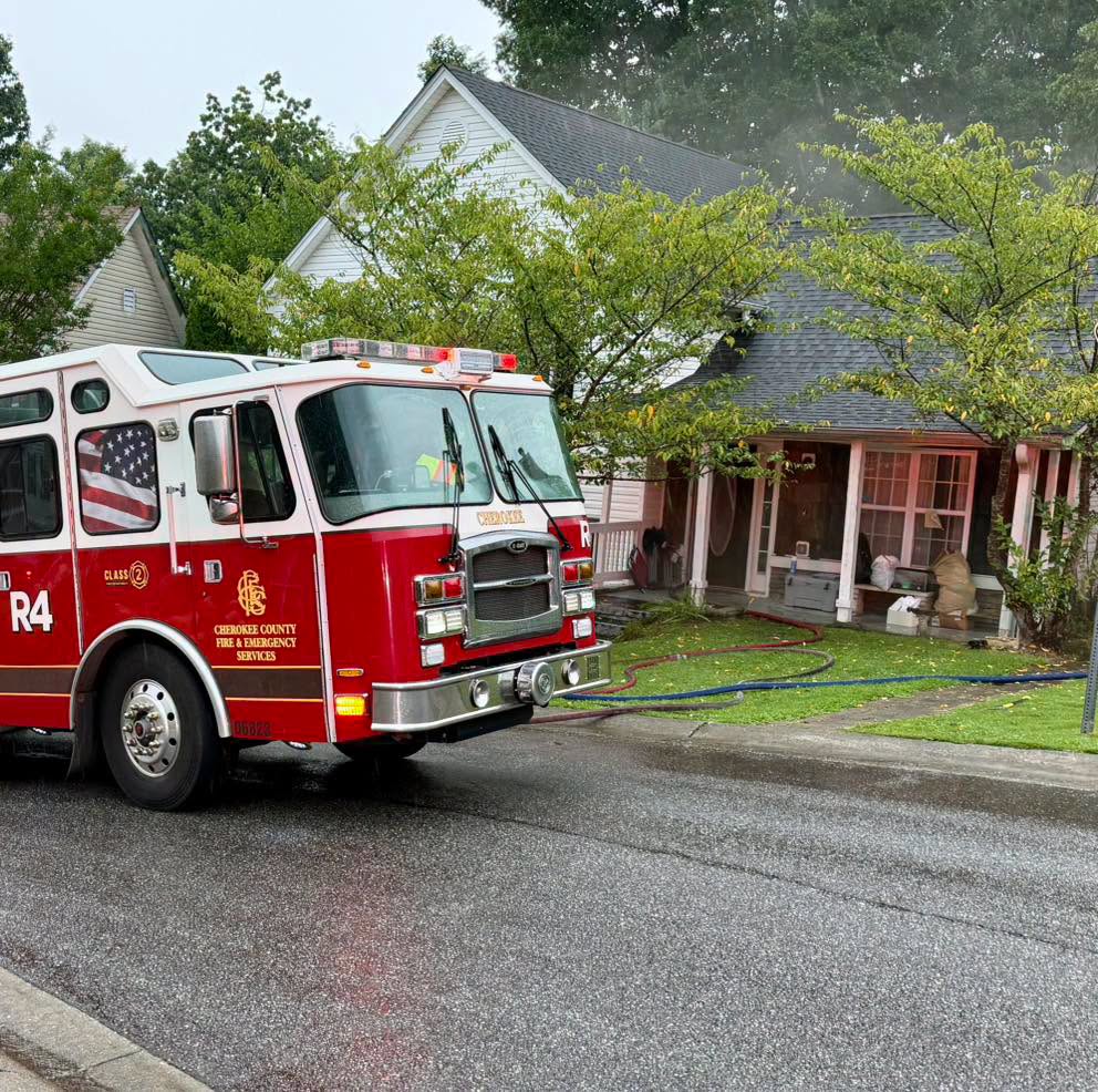 Cherokee County fire engine parked in front of a two‑story house with hoses on the wet ground and light smoke near the roof.